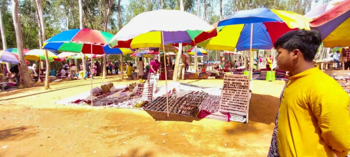 Colorful market scene with vibrant umbrellas shading various stalls. Displays of jewelry and trinkets are visible. People browse through the market, walking on the sandy ground. Trees in the background provide a natural setting. A person stands in the foreground.