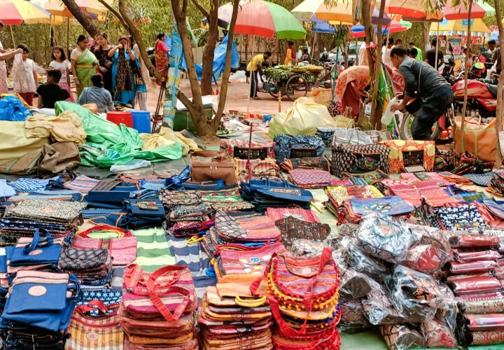 A colorful outdoor market displays a variety of bags and textiles on the ground. Shoppers and vendors are seen under umbrellas, with trees and other market stalls in the background.