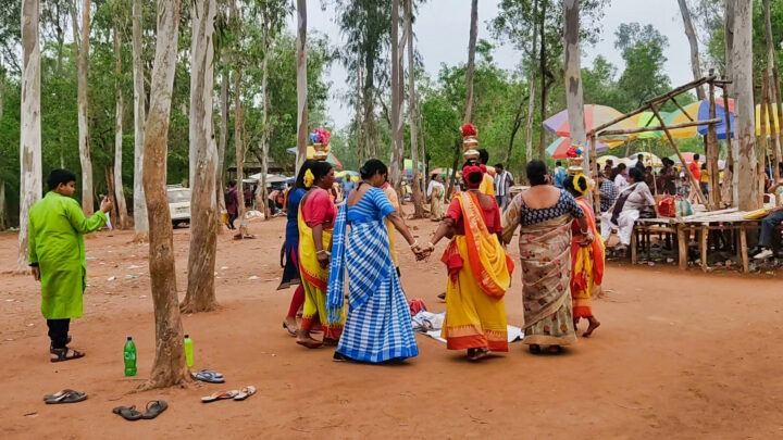 A group of women dressed in colorful traditional attire walk in a circle, carrying pots on their heads in a forested area. People are gathered around and stalls are visible in the background.
