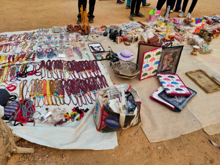 Various handmade crafts, including bead necklaces, bracelets, woven baskets, and colorful paintings, are displayed on cloth sheets on the ground. A bag filled with items and people’s feet are visible in the background.