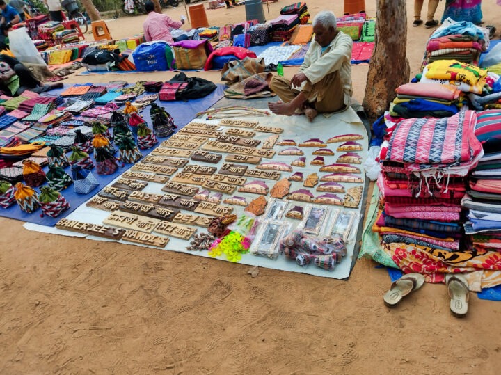 A street vendor sits on the ground beside a display of handcrafted wooden signs, colorful patterned bags, and neatly stacked textiles. The items are spread out on a tarp at an outdoor market.