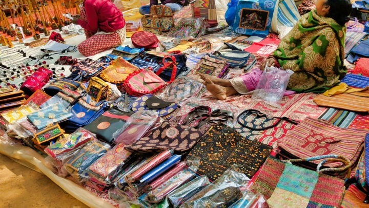 A market stall displays a variety of colorful handmade bags, wallets, and textiles. Two vendors are seated behind the items, with other goods and crafts visible in the background. The scene is vibrant and busy with intricate patterns.