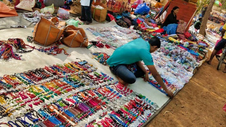 A vendor in a turquoise shirt is arranging colorful handmade jewelry on display at an outdoor market. Various bags and goods are spread out on a large cloth, with more items visible in the background.