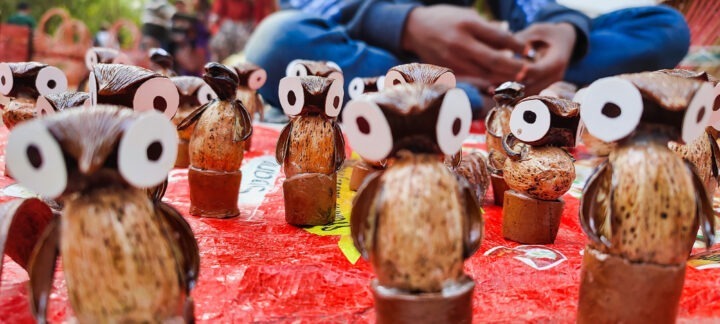 Close-up of handmade owl figurines made from natural materials, such as seeds and nuts, arranged on a vibrant red cloth. The owls have large white eyes. A person in the background is partially visible, seated and seemingly crafting.
