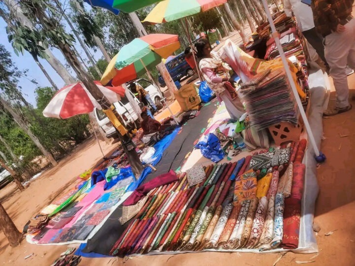 An outdoor market stall displays colorful fabrics arranged on the ground under umbrellas. Several people are browsing or selling. The setting is surrounded by trees and parked vehicles.
