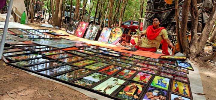 A person sits on the ground surrounded by various framed artworks displayed for sale in a wooded outdoor setting. The artworks feature colorful and diverse designs. There is a red car visible in the background.