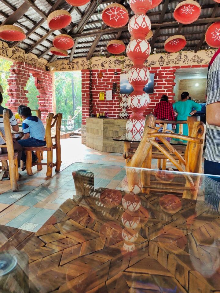A colorful, rustic indoor cafe with wooden furniture and vibrant decor. Red lanterns hang from the ceiling, and walls feature red brick and painted patterns. People are seated at tables, with natural light streaming in from large windows.
