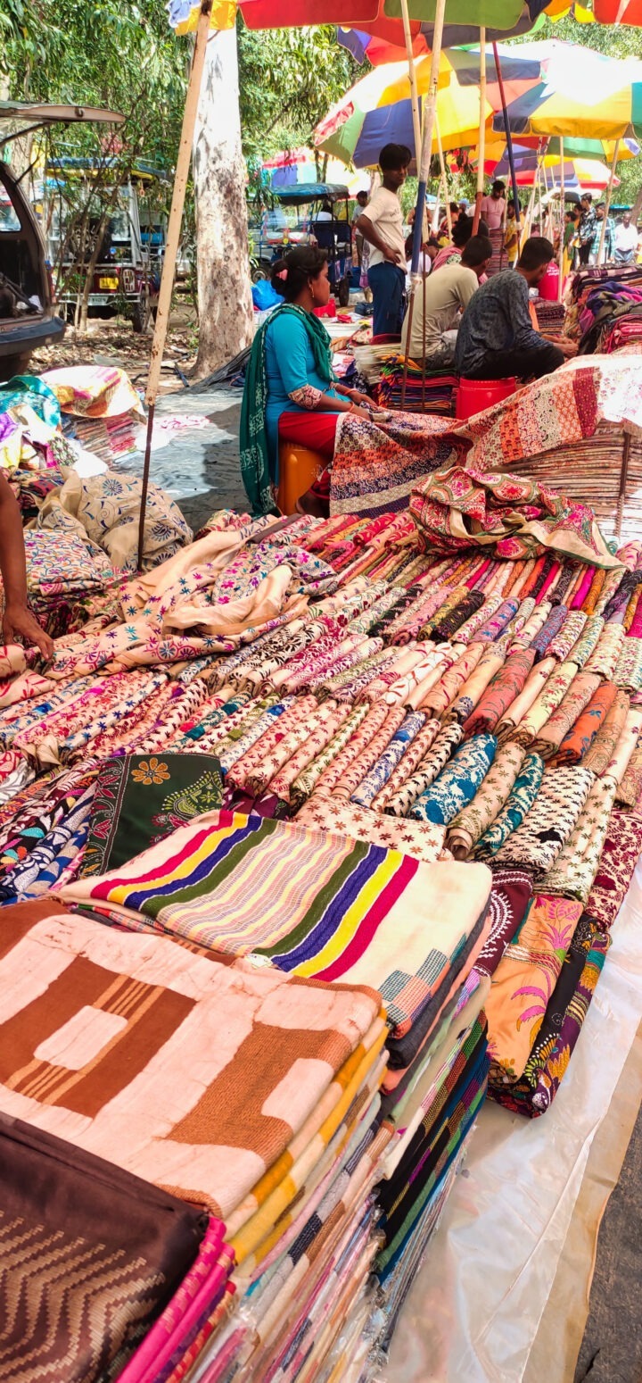 A market stall displays a variety of colorful fabrics and quilts. Patterns include stripes, florals, and geometric designs. Several people browse items under large umbrellas, with more market activity visible in the background.