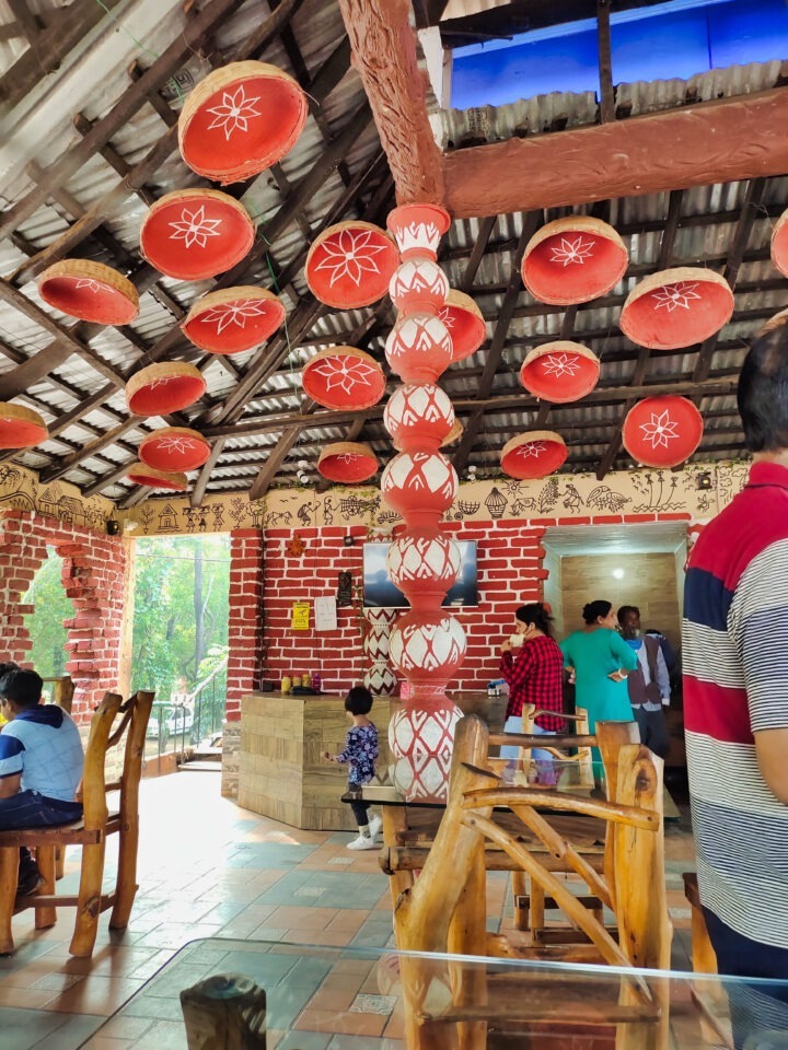 A group of people sit in a rustic restaurant with wooden furniture and decorative red pots hanging from the ceiling. The walls are adorned with tribal art. A child stands near a painted pillar in the center, and a TV is mounted on the wall.