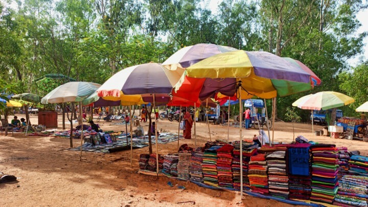 Market scene with colorful umbrellas providing shade over various textiles and fabrics displayed on tables and racks. Surrounding trees and a clear sky create an open, outdoor setting. People can be seen browsing and interacting in the background.