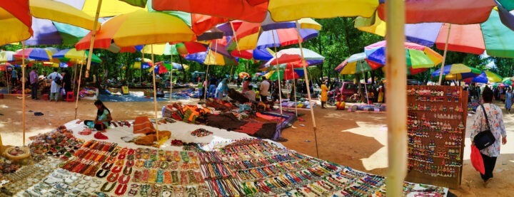 A busy outdoor market with colorful umbrellas providing shade. Stalls display a variety of items including jewelry, souvenirs, and textiles. Shoppers and vendors interact amidst the lively setting, surrounded by trees.