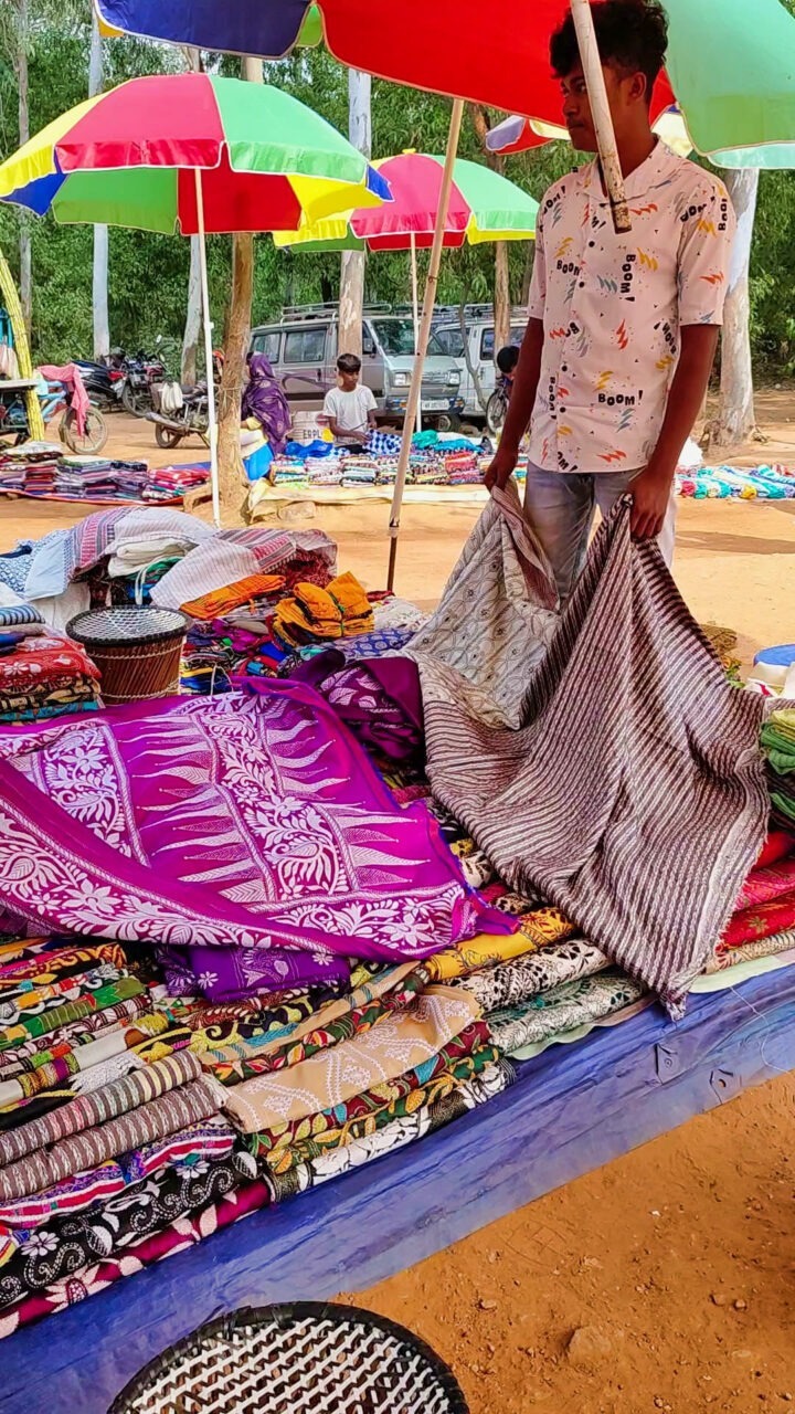 A vendor at an outdoor market is arranging colorful, patterned textiles on a table. The textiles vary in colors and designs, including florals and geometric patterns. Bright umbrellas shade the area, and other stalls and trees are visible in the background.