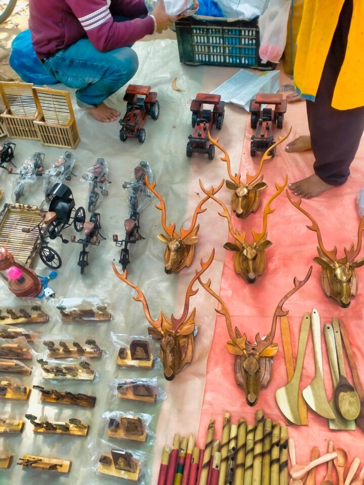 A market stall displays various handcrafted wooden items, including deer head ornaments, cutlery, toy trucks, bamboo flutes, and small cages. Two people are arranging the items on a beige and red cloth.