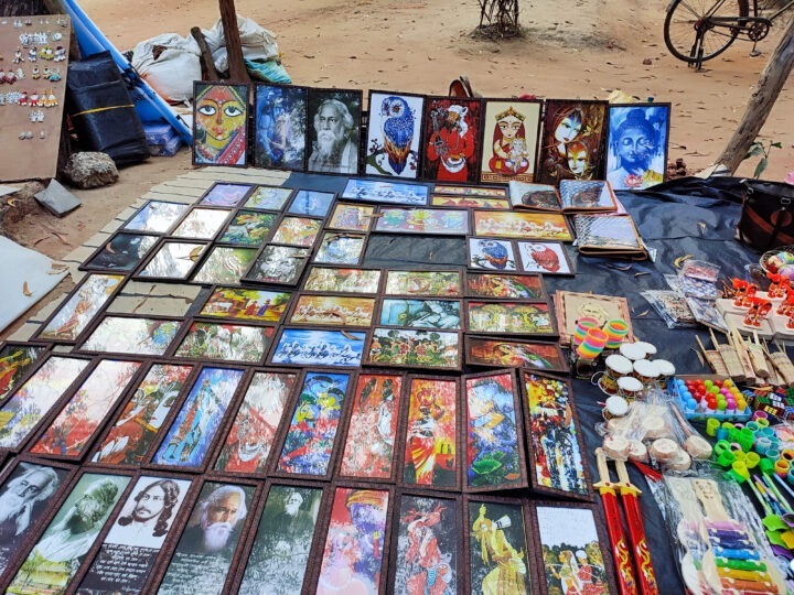 A market stall displays various colorful framed artworks laid out on a tarp. The art includes portraits, landscapes, and abstract designs. Some small toys and crafts are also visible. A bicycle is partly seen in the background.
