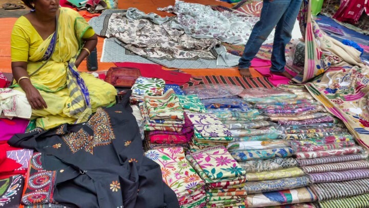 A vendor displaying a variety of colorful fabrics and garments on a marketplace floor. The assortment includes embroidered and patterned textiles. A seated vendor in a yellow saree is visible on the left, with more fabrics spread out nearby.