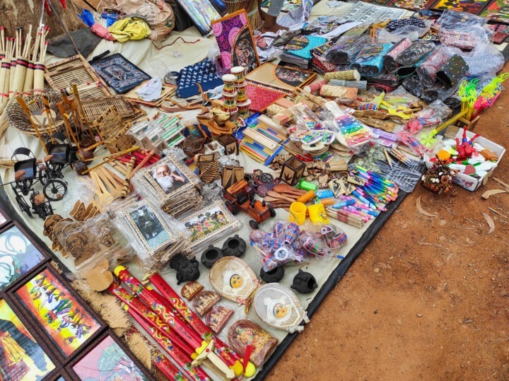 A colorful market stall displays a variety of handcrafted items, including wooden toys, picture frames, painted fans, masks, woven baskets, and assorted souvenirs. Items are arranged on a cloth spread over the ground.