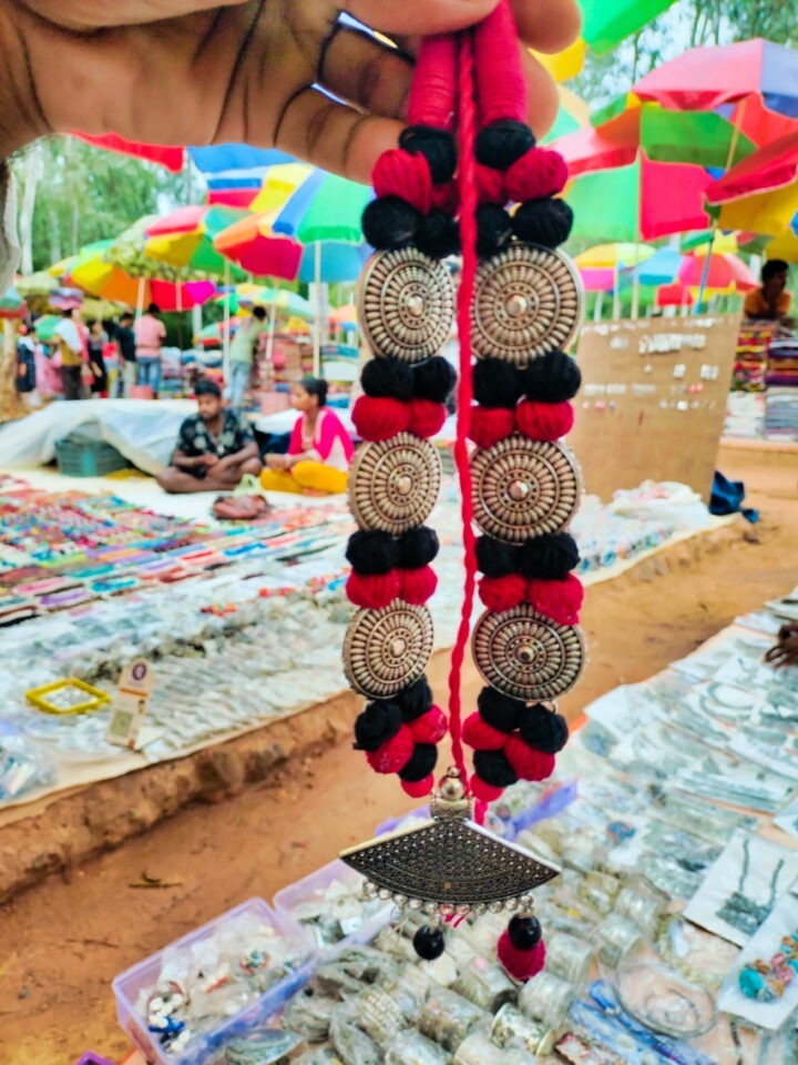 A hand holding a traditional silver and red beaded necklace in an outdoor market. The background features colorful umbrellas and various jewelry displayed on tables.