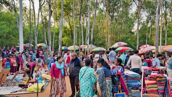 A bustling outdoor market scene with numerous people shopping and socializing. Colorful umbrellas shade various stalls displaying goods. Tall trees surround the area, adding a natural backdrop.