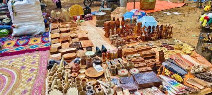 A market stall displays an array of wooden crafts, including carved figures, bowls, boxes, and other items. There are also textiles and small trinkets. The ground is covered with patterned cloths and tarps.