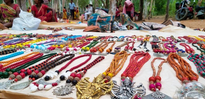 A display of colorful beaded necklaces and metal pendants laid out on a white cloth at an outdoor market. Shoppers and vendors are in the background, with trees surrounding the area.