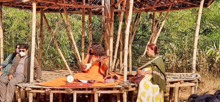 A musician in an orange outfit sits cross-legged playing an instrument in a bamboo gazebo. Two people sit beside, one wearing a mask and sunglasses, the other in a green shawl and floral dress. Trees are visible in the background.