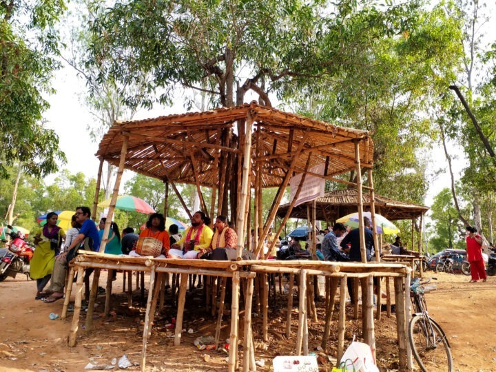 A rustic wooden structure with a thatched roof serves as a shaded seating area. Several people are seated inside, engaged in conversation. Bicycles are parked nearby, and trees surround the spot, casting shade.