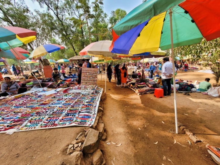 A vibrant outdoor market scene with colorful umbrellas shading various stalls. Tables display an array of beaded jewelry and crafts. People are browsing the items while others are seated nearby, surrounded by trees and a clear blue sky.