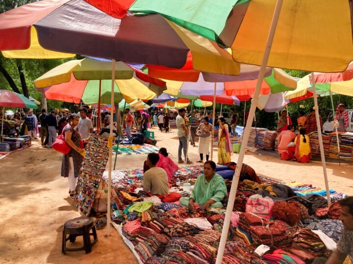 A bustling outdoor market with colorful umbrellas providing shade. Vendors display a variety of textiles and accessories on the ground. Shoppers browse the offerings, and the scene is lively with people interacting and exploring.