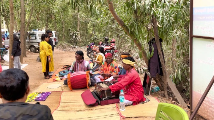 Musicians are sitting on a mat in a wooded area, playing traditional instruments. People are standing and sitting nearby. A parked car and motorcycles are visible in the background.