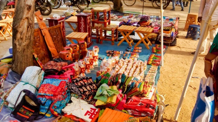 A vibrant market stall displays a variety of colorful textiles, handmade wooden crafts such as chairs and decorative owls, and other small items. Bicycles are visible in the background, and the setup is shaded by a tree.