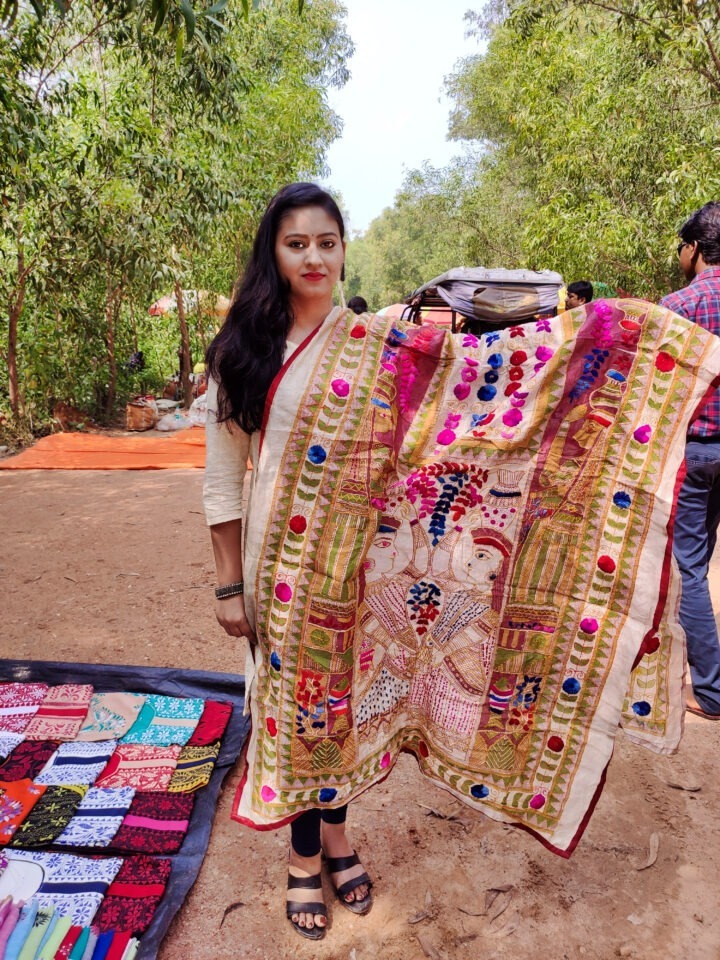 A woman stands on a dirt path in a forested area, holding a colorful embroidered textile featuring intricate designs. Nearby, a display of folded textiles is visible on the ground. Other people can be seen in the background.