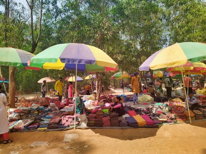 A vibrant outdoor market scene with colorful umbrellas providing shade over stalls displaying various fabrics. People are browsing and interacting in the background. Tall trees surround the market area, casting shadows on the ground.