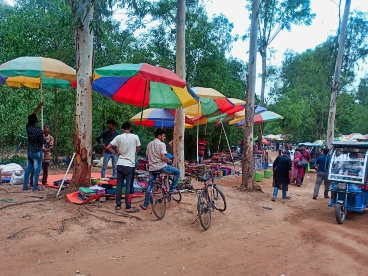 A market scene with colorful umbrellas covering various stalls set up along a dirt road. People are browsing the merchandise, and bicycles are parked nearby. Trees surround the area, and a three-wheeled vehicle is parked on the right.