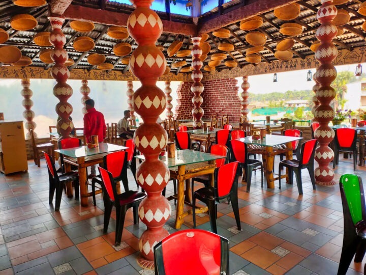 A restaurant with an open-air feel features red and black chairs around square tables. The space is decorated with ornate red and white pillars, a tiled floor, and a ceiling adorned with woven baskets. One person is visible in the background.