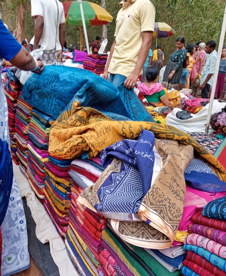 A market scene with people browsing and arranging colorful folded fabrics displayed on tables. Various patterns and colors are visible. In the background, people and trees can be seen under vibrant umbrellas.