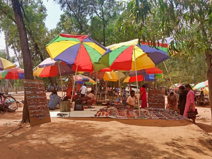 A market scene with colorful umbrellas providing shade over several stalls. Vendors display various items, such as jewelry and crafts, on wooden tables and stands. Shoppers browse the goods on a dirt path surrounded by trees.