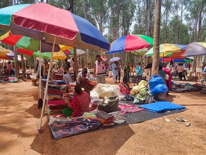 Outdoor market scene under colorful umbrellas. Vendors display textiles and various goods on the ground. Trees surround the market, and shoppers browse the items. Bright and busy atmosphere with natural light filtering through the trees.