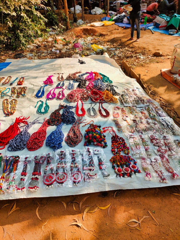 A display of colorful handmade jewelry items, including necklaces, earrings, and bracelets, arranged on a white cloth spread on the ground at an outdoor market. The setting includes surrounding foliage and a dirt pathway.