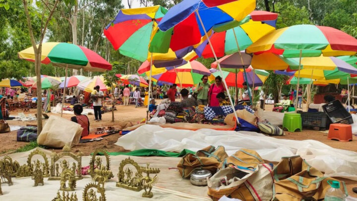 A vibrant outdoor market scene with multicolored umbrellas providing shade. Various goods, including metal figurines and bags, are displayed on the ground. People are browsing and interacting among the stalls amidst trees in the background.